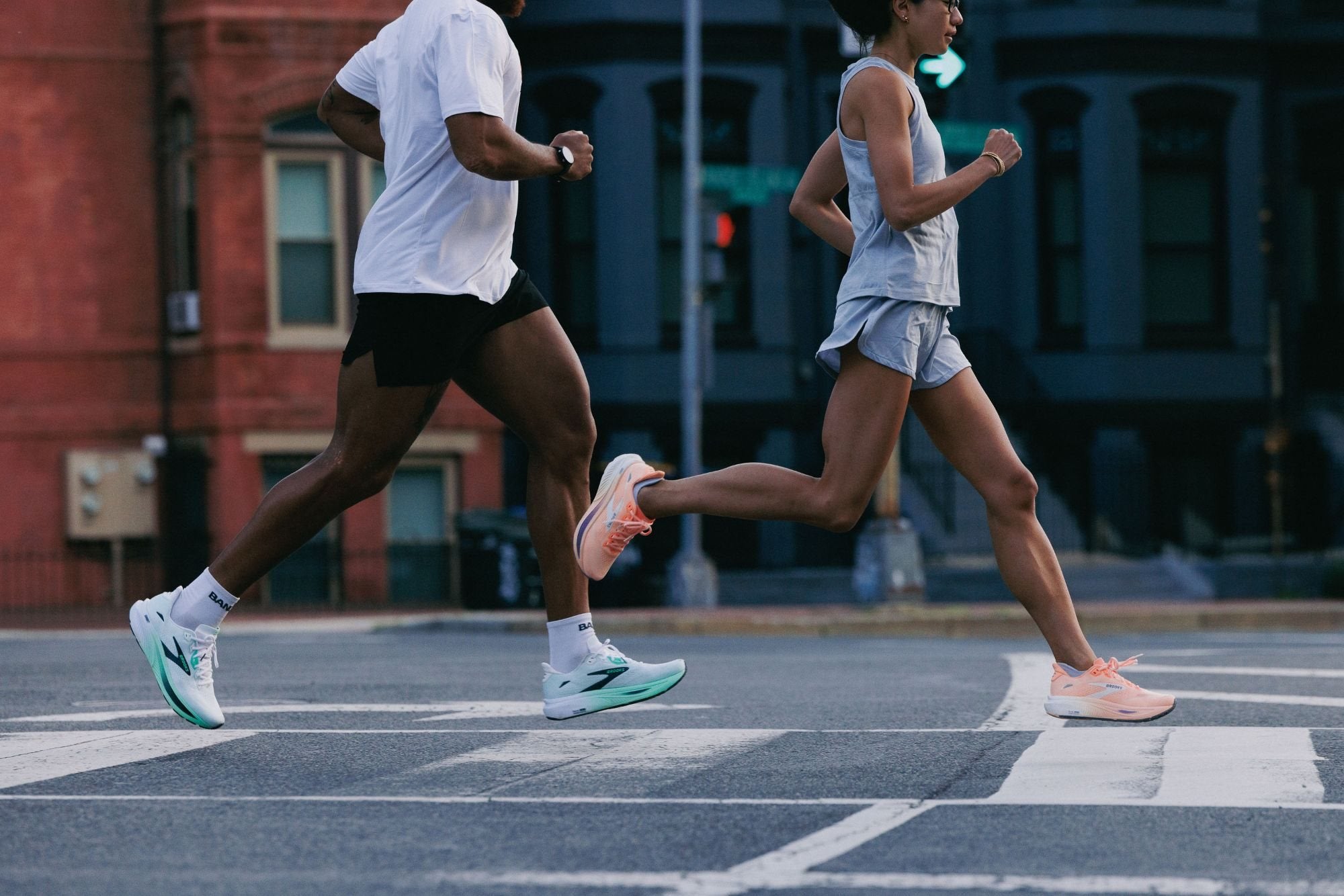 Two runners in running shoes stride across a city street crosswalk, one in a white shirt and black shorts and one in a light grey tank and shorts, with a brick row house behind them.